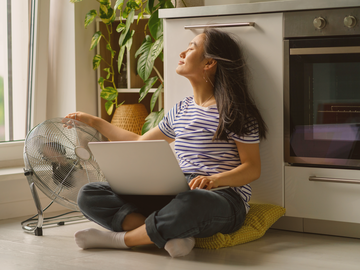 Frau sitzt am Laptop auf dem Boden in der Kueche neben einem Ventilator